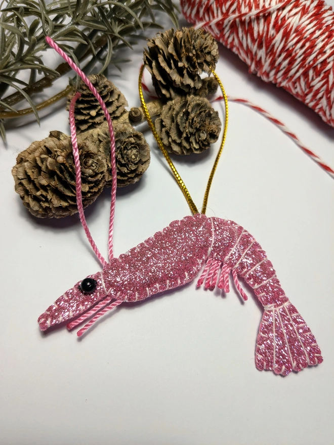 Hand-stitched pink glitter King Prawn Christmas decoration with pink twine antennae and legs, black beady eyes and gold hanging loop, nestled amongst festive greenery and for cones.