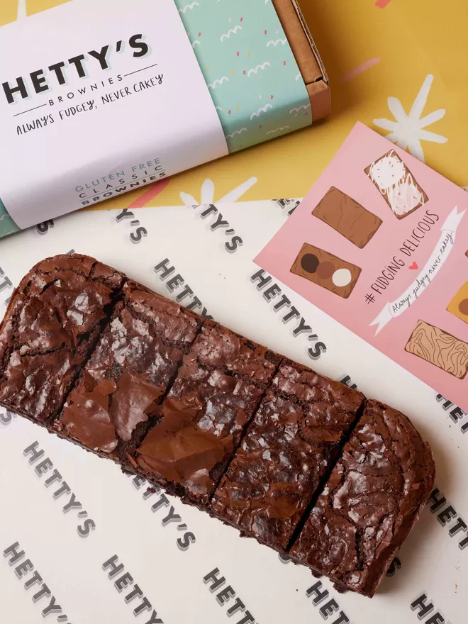 Five slices of gluten free classic fudge brownie in a flat lay with branded box and postcard