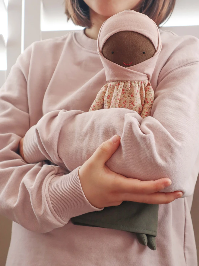 Handmade cloth doll wearing a blush pink hijab, a beige floral long-sleeve blouse, and an olive green skirt. The doll has dark fabric skin, simple stitched eyes and a small red smile, with a BOKUNO label on the side. 