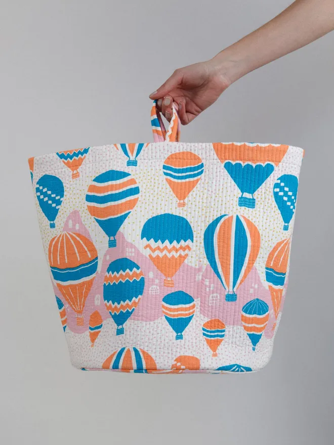 Balloons at Dawn Quilted Storage Basket, a person holding an orange and blue hot air balloon pattern basket against a grey backdrop. 