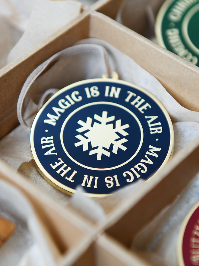 A navy blue and gold enamel Christmas decoration, with the words Magic Is In The Air surrounding a gold snowflake, is tucked into a sectioned box.