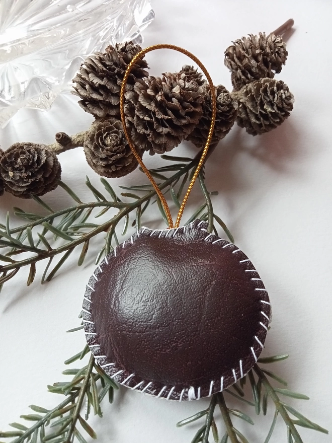 Hand stitched leatherette Christmas pudding decoration placed on a branch of small pine cones, to show it's plain brown reverse