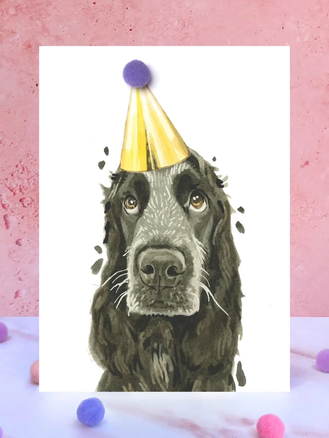 A greeting card featuring a hand painted design of a black/blue roan Cocker Spaniel, stood upright on a marble surface surrounded by pompoms. 