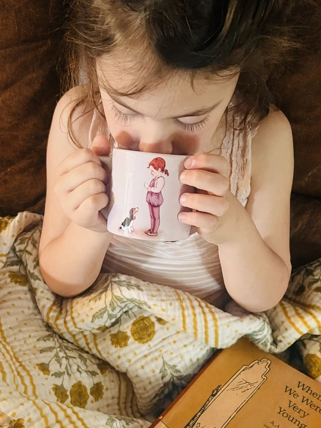 a girl drinking her hot drink from a small ceramic mug featuring a story book style illustration of a little girl and her beagle puppy