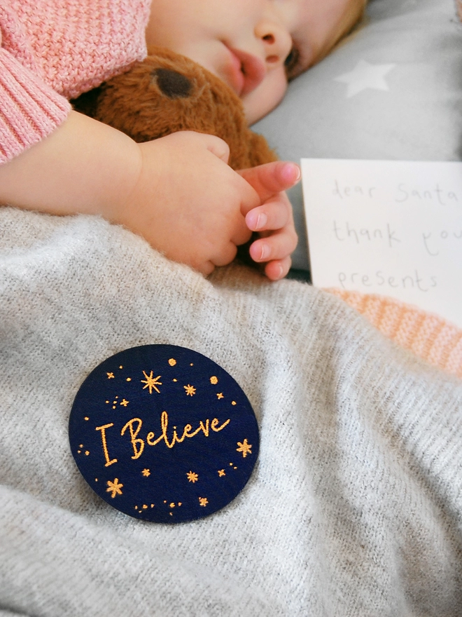A navy blue woven patch with golden stars and the words "I Believe" is attached to a grey blanket, which lays over a sleeping toddler.