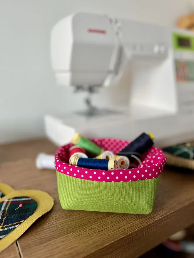 Small green fabric storage basket holding bright cotton reels, on a table beside a sewing machine.