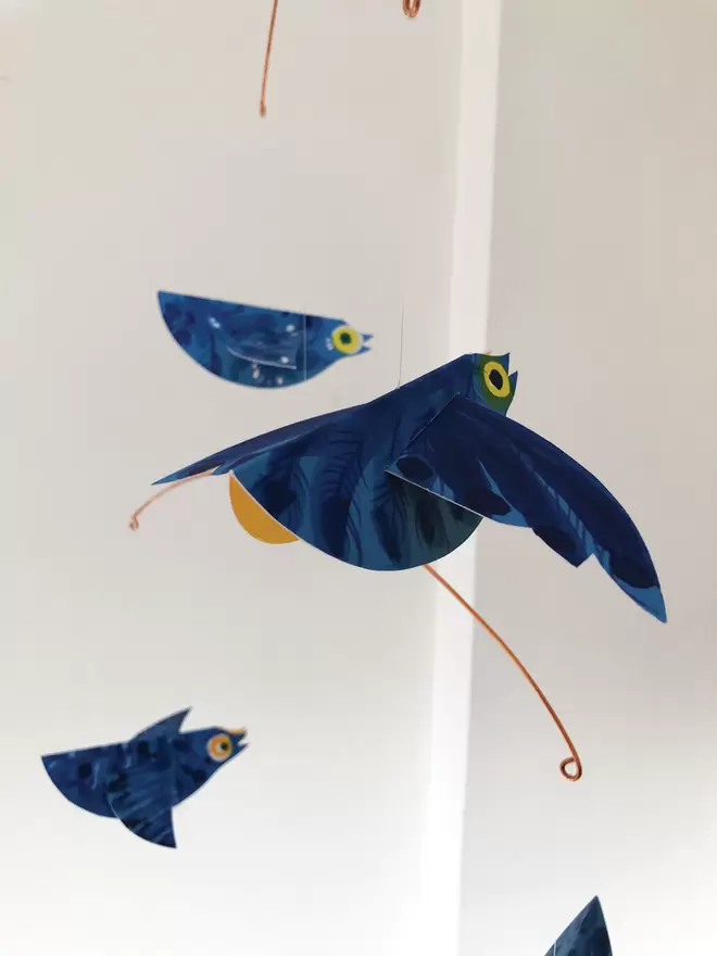 Three bright blue patterned paper birds mobile hanging against a white wall. They show sunshine-yellow bellies.
