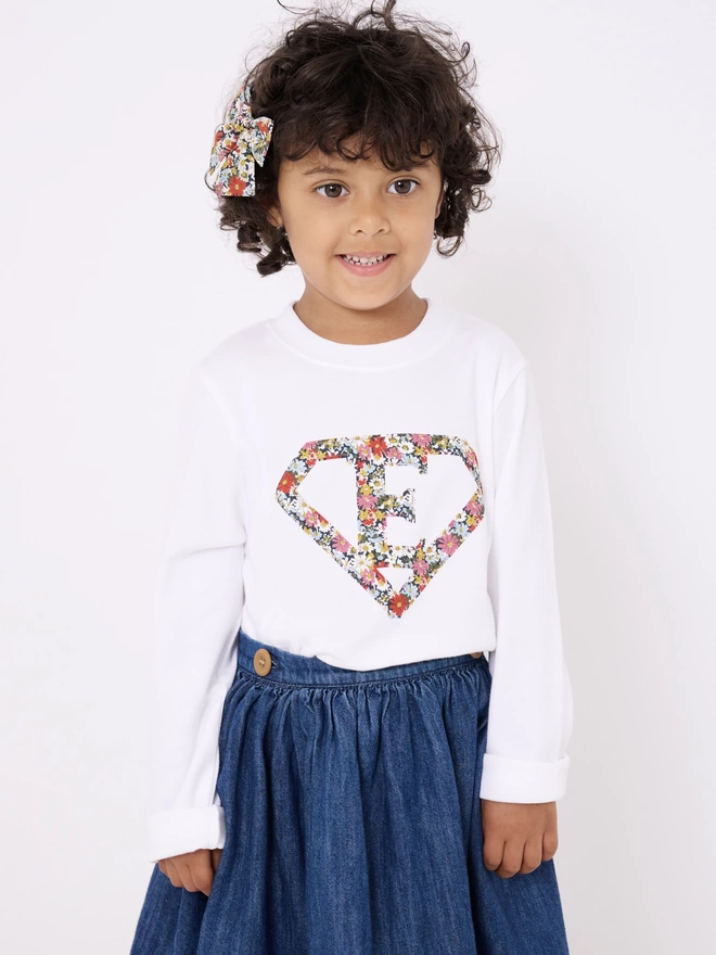 A girl wearing a white long sleeve t-shirt appliquéd with a superhero motif  featuring a letter E initial inside it. The appliqué is made from a floral Liberty print .