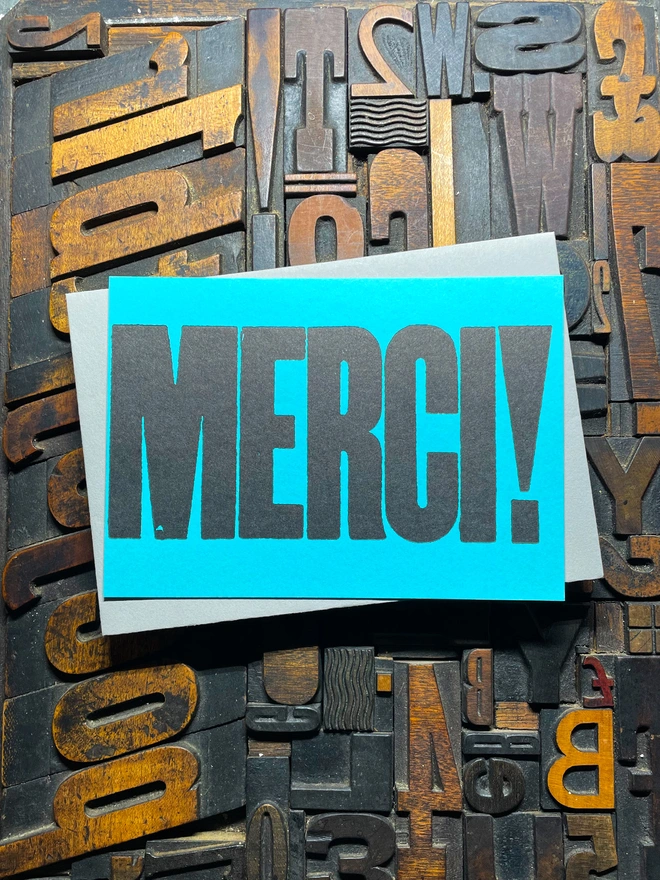 A bright turquoise thick duplex card with the word "MERCI!" printed in bold, black letters lies on a surface covered with various letterpress blocks.