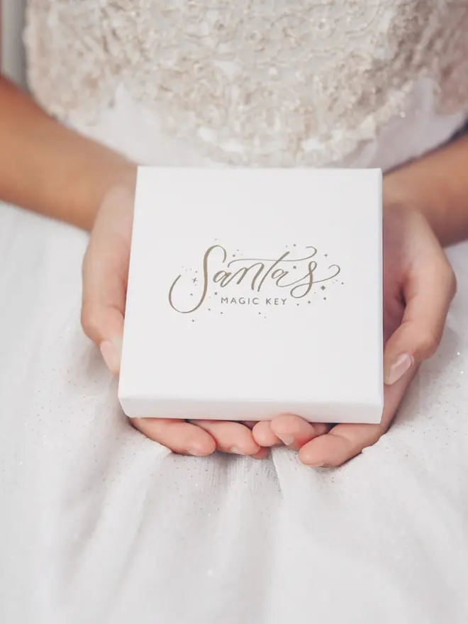 A child holds a white gift box in both hands. The lid has the "Santa's Magic Key" logo in gold foil.