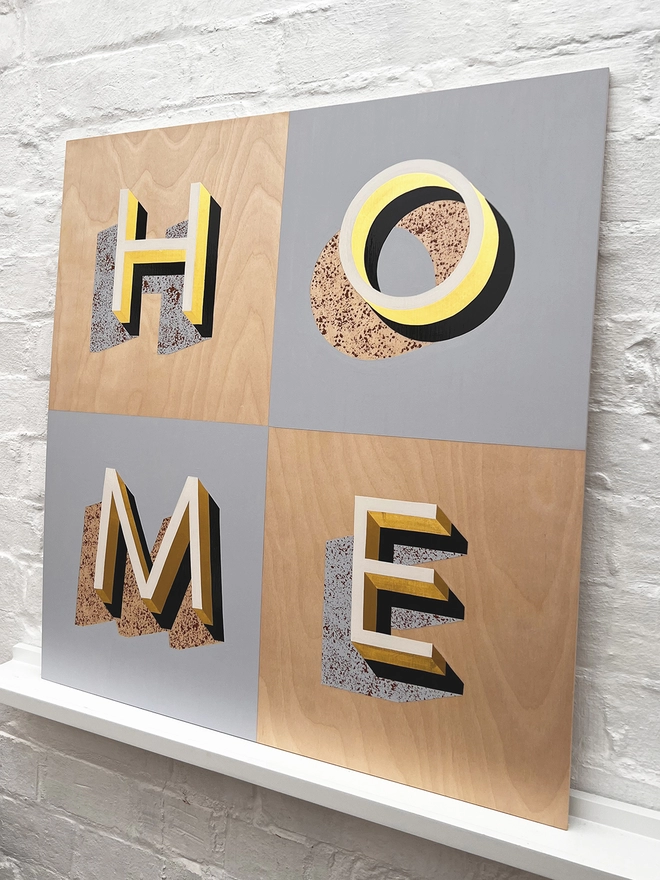 HOME hand painted sign in 23 carat gold leaf, pale blue, off white, tan, against a white brick wall, at an angle. 