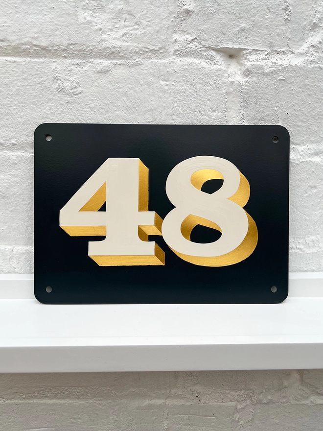 Hand painted house number against a white brick wall, number 48 in off white and directionally burnished 23 carat gold leaf, on anthracite grey background. 
