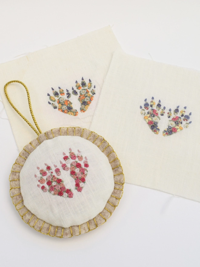 Showing a pair of rabbit feet in each of the 3 embroidered designs.  Sunshine Garden & Floral Meadow on the plain linen squares.  The Pink Roses design is shown as a felt bauble.