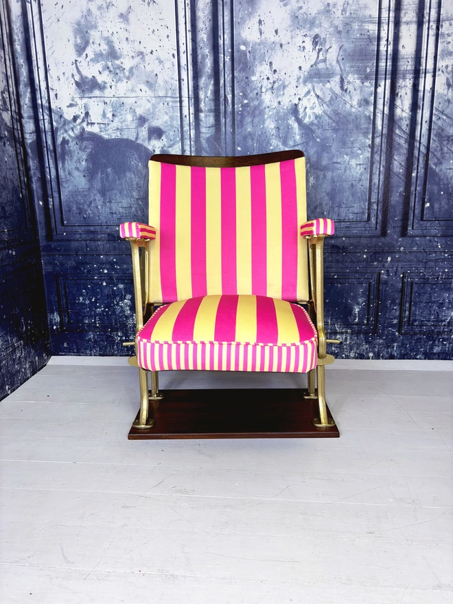A pink and yellow vintage cinema seat.  The cinema seat has gold legs and is mounted onto a wooden plinth.  The fabric is pink and yellow striped.