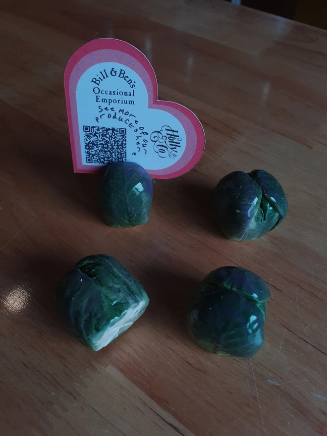 A set of four handmade ceramic Brussels sprout name card holders arranged on a wooden surface. Each glossy green sprout has individually modelled leafy details, and one is shown holding a heart-shaped display card.