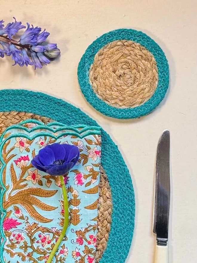 Seagrass coaster with teal rings, shown on a light surface with purple flowers. 