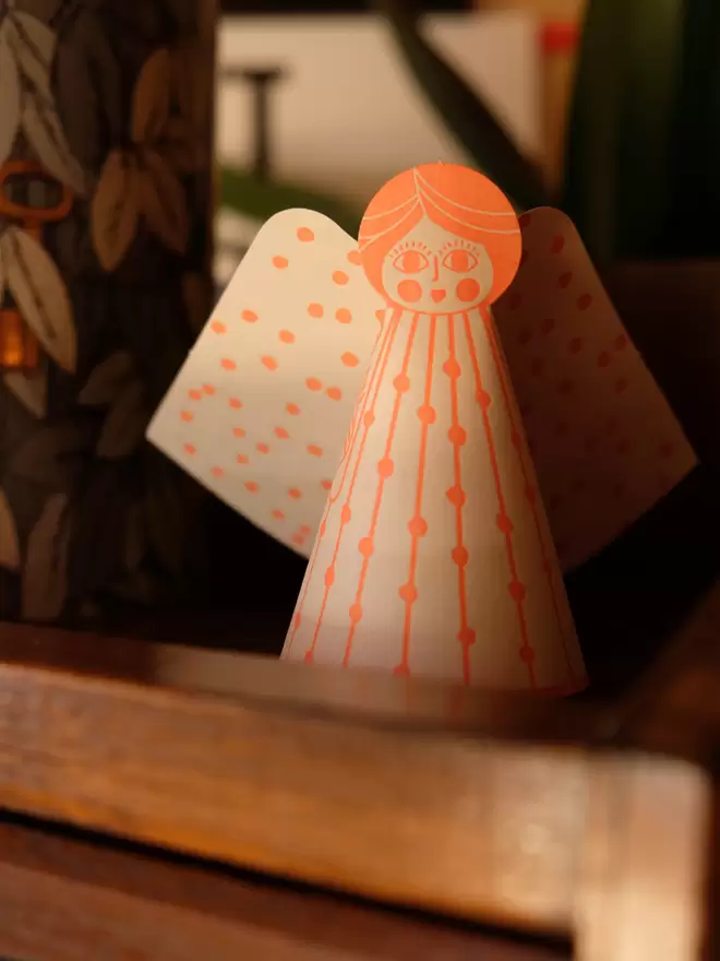 Orange 3D angel card standing on cabinet in dusky light.