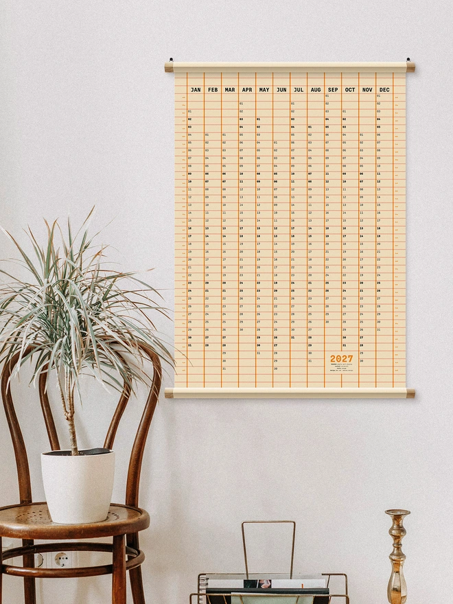 2027 Wall Planner (Squared) in orange on white wall, plant to the side.