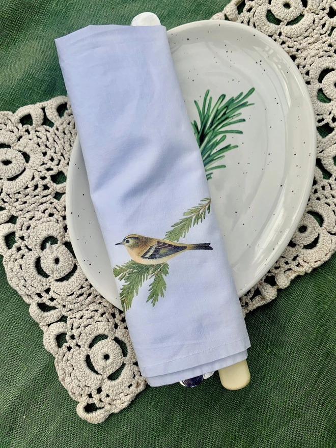 A white cotton napkin printed with a vintage illustration of a goldcrest, on an oval side plate and vintage crochet lace table runner