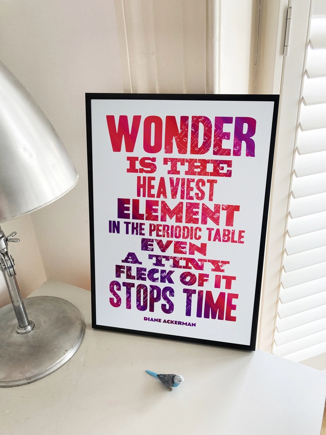 Framed multicoloured typographic print of “Wonder is the heaviest element in the periodic table, even a tiny fleck of it stops time” by Diane Ackerman. The print rests on a grey desk next to a lamp with a small blue budgie.