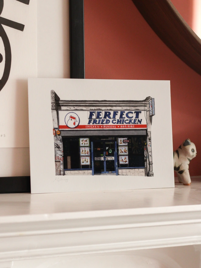 An architectural illustration of the Ferfect Fried Chicken storefront on a print, displayed on a white shelf alongside small cat and elephant figurines.