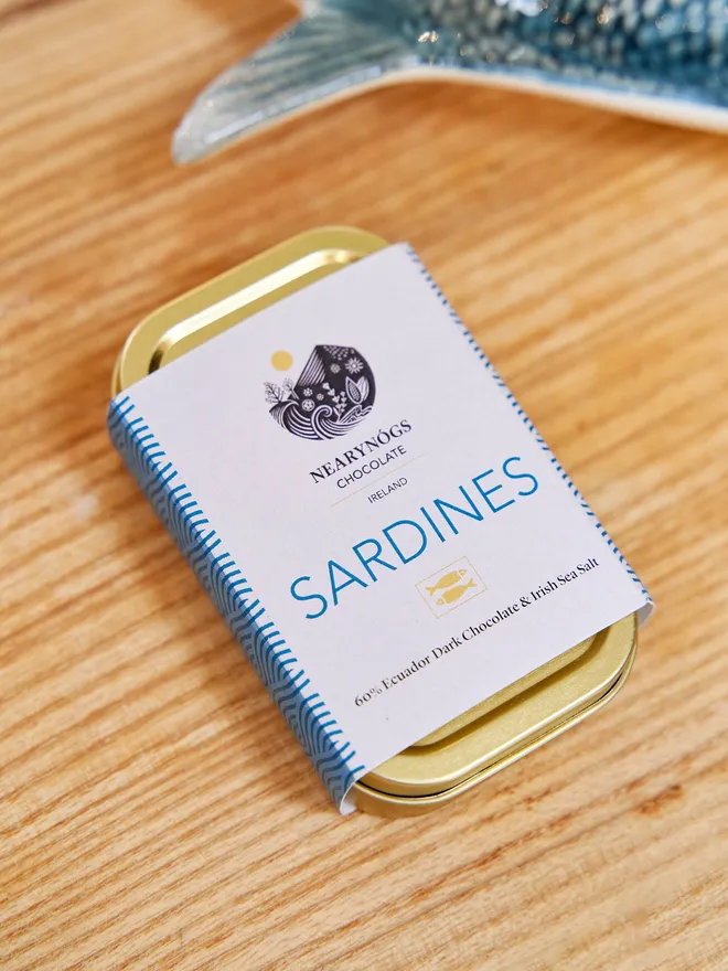 Sardines Dark Chocolate 60% (Tin Of 4), a tin of chocolate fish sitting on a wooden surface. 