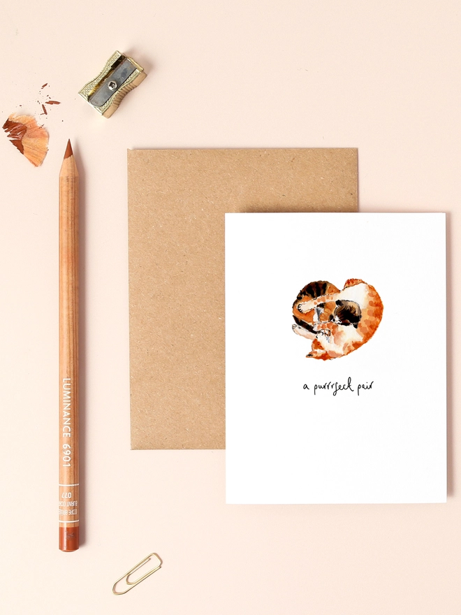 An image of a mini, rectangular white greetings card with an illustration of a couple of hugging ginger cats shaped in a heart. A Purrrfect pair hand written message is seen under the central illustration.