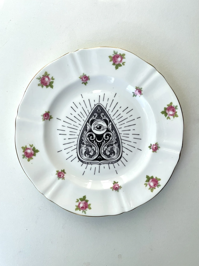 vintage plate with an ornate border, with a printed vintage illustration of a Ouija Planchette in the middle 