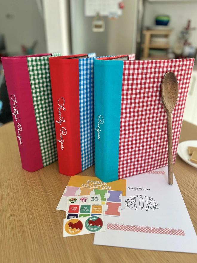 3 gingham recipe binders in red, blue and green, standing on a kitchen table with recipe template pages and stickers beside it. Fresh baking in background.