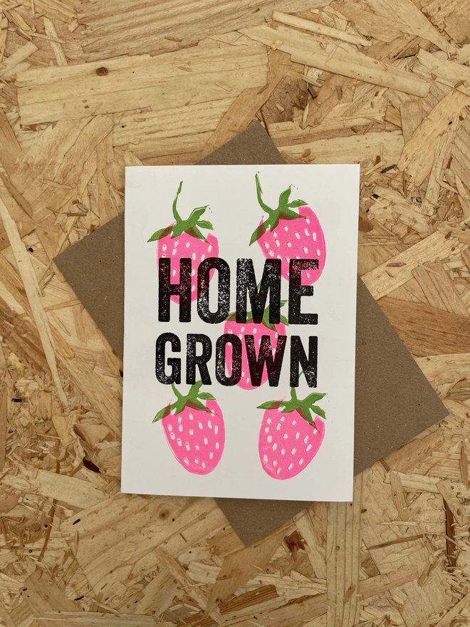 A white greeting card on a wooden surface shows  illustrated Pink strawberries with green leaves surrounding bold black text that reads “HOME GROWN.”