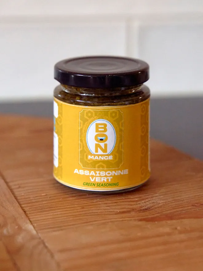 Bon Mange Green Seasoning in a jar with a yellow label on a wooden board atop a black worktop with a white tile background