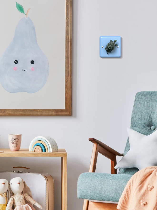 A tortoise light switch by Candy Queen Designs on a white wall next to an art print of a happy blue smiling pear in a brown wooden frame and a blue armchair with wooden arms. There is a wooden shelf table to the left with a wooden rainbow toy and a mug sitting on it and two soft toy rabbits sitting in front of it. The light switch is a blue nursery dimmer light switch with a grey tortoise as the rotary knob to turn the lights on and off. The light switch plate is pale pastel blue and made of metal, epoxy coated steel by Varilight. The tortoise is made of plastic. The animal light switch brand is Candy Queen Designs.