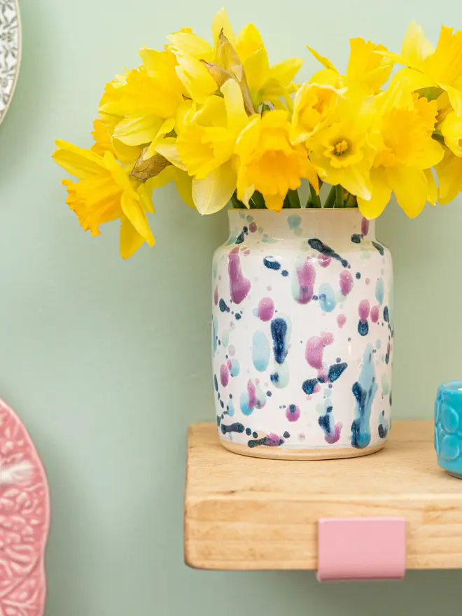 Sweetpea Medium Vase, a colourful vase with a collection of yellow flowers in it. It sits in front of a green backdrop on a wooden surface. 