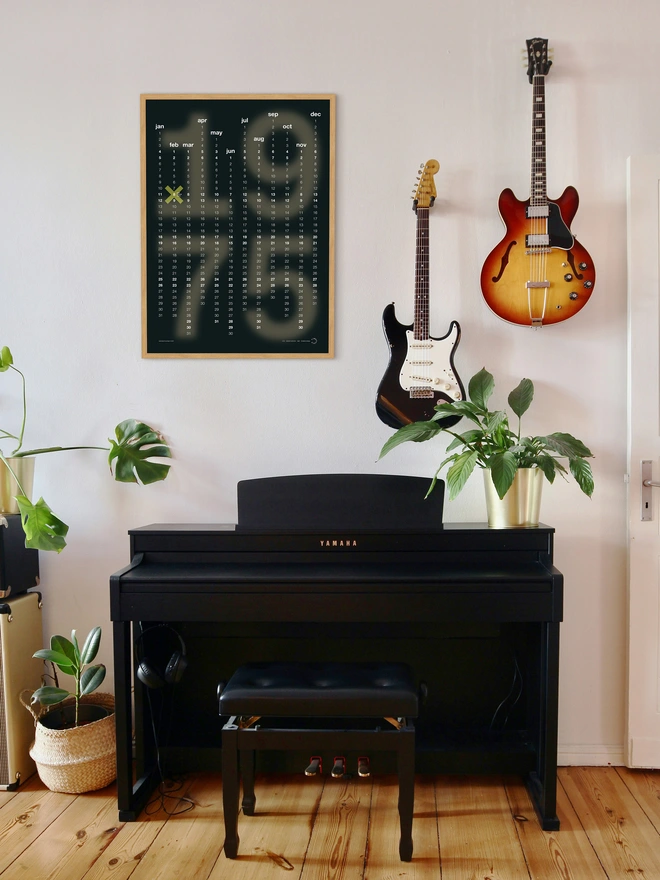 Framed Black Minx Personalised Date Poster on white wall, next to a guitar, a plant and piano