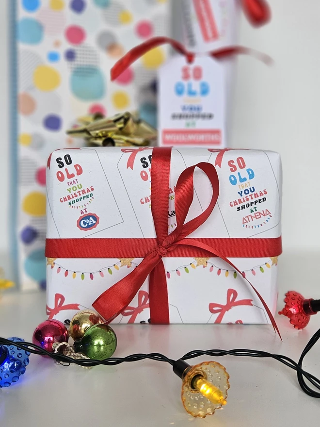  A rectangular present gift wrapped in a design that says so old that you Christmas shopped at Woolworths, C&A, Sock Shop, Tie Rack, Athena on an illustrated white tag with a red bow. The text is written in brightly colours with a string of coloured fairy lights.