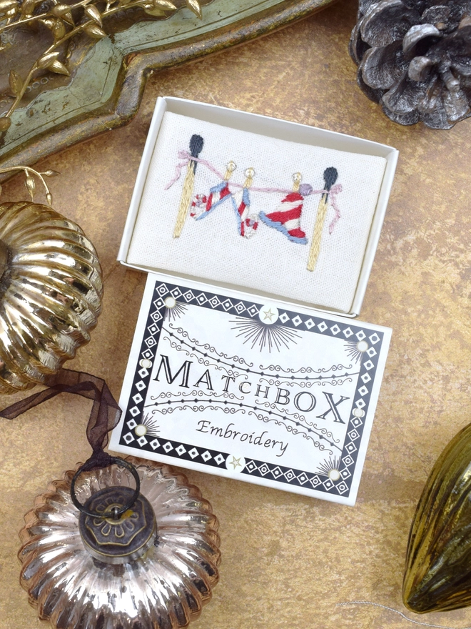 The elf laundry embroidery, inside a Matchbox displayed on a gold background with glass baubles.