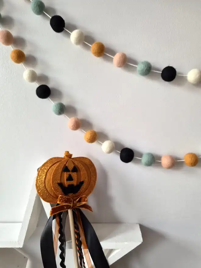 A pumpkin wand, with a felt ball garland hanging above in soft, Halloween shades.