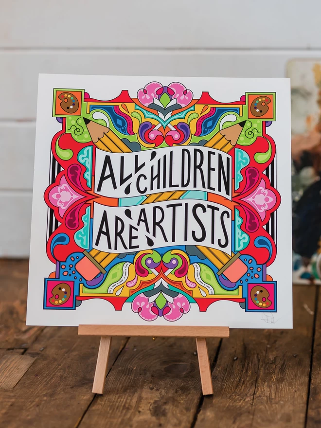The print is on a small wooden easel, sitting on floorboards in front of a white brick wall. The words All Children are Artist sit at the centre of this multi-coloured vibrant illustration which includes a cross of pencils, and artists palettes the corners. 