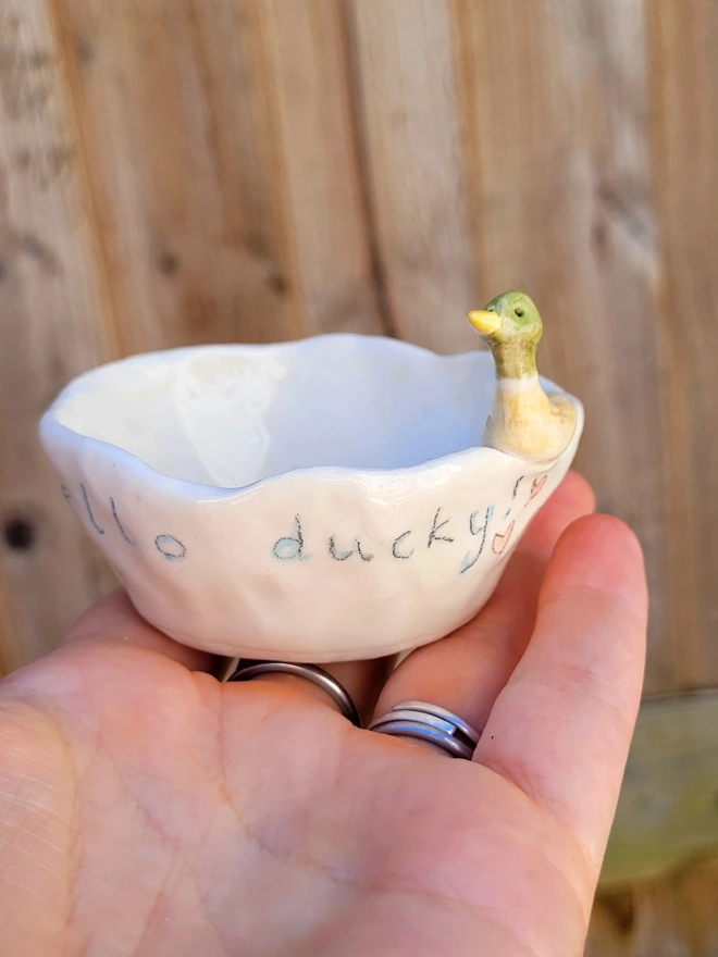 small ceramic ivory pot held in a hand with a modelled drake duck on the rim and the words hello ducky on the pot