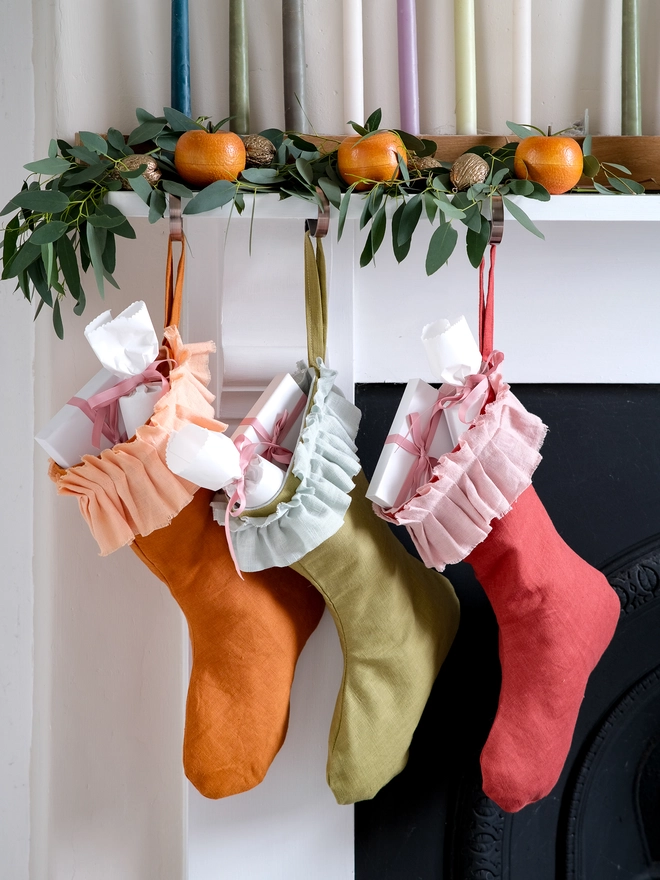 handmade linen Christmas stockings in warm orange, olive green and red hang beneath a festive mantel, each finished with a soft, gathered ruffle cuff.