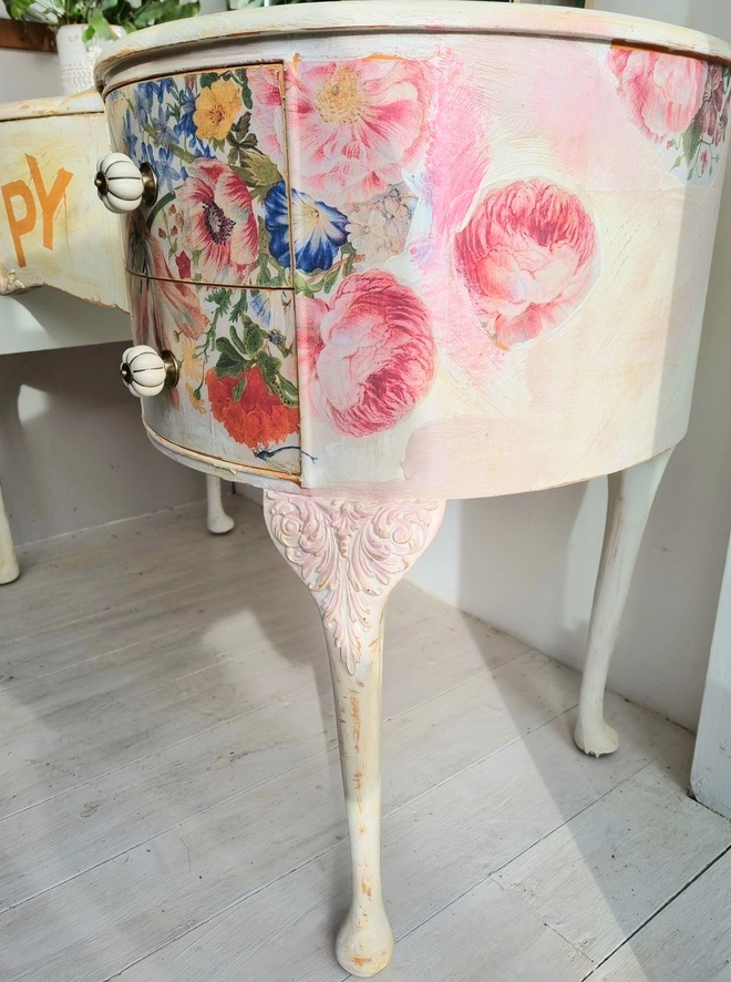 An antique kidney-shaped dressing table with chair and a trio of mirrors. Upcycled in cream and pale pink with decoupage florals in oranges and pinks. Text across the table read ‘flowers make me happy’ in the orange of the undercoat.