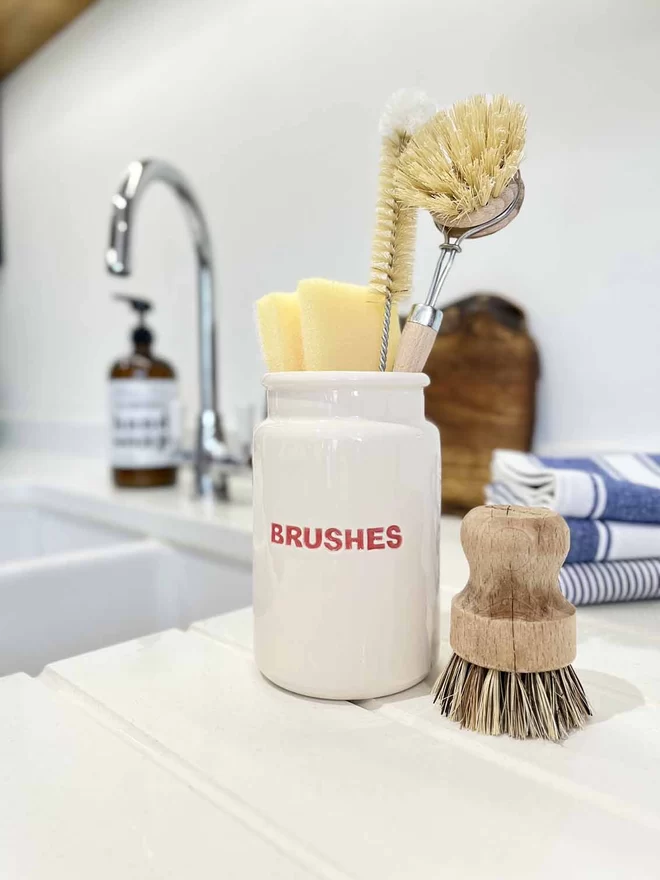 A handmade ceramic jar stands by a kitchen sink with the word ‘brushes’ hand painted in red.