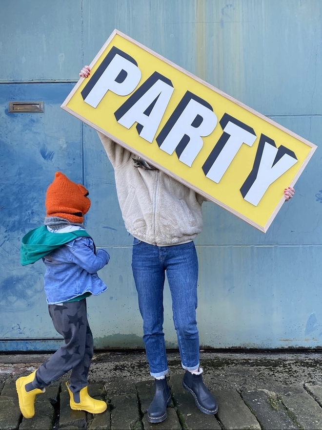 a person holding a painted wooden sign above their head which reads PARTY on a yellow background. With a child standing next to her.  