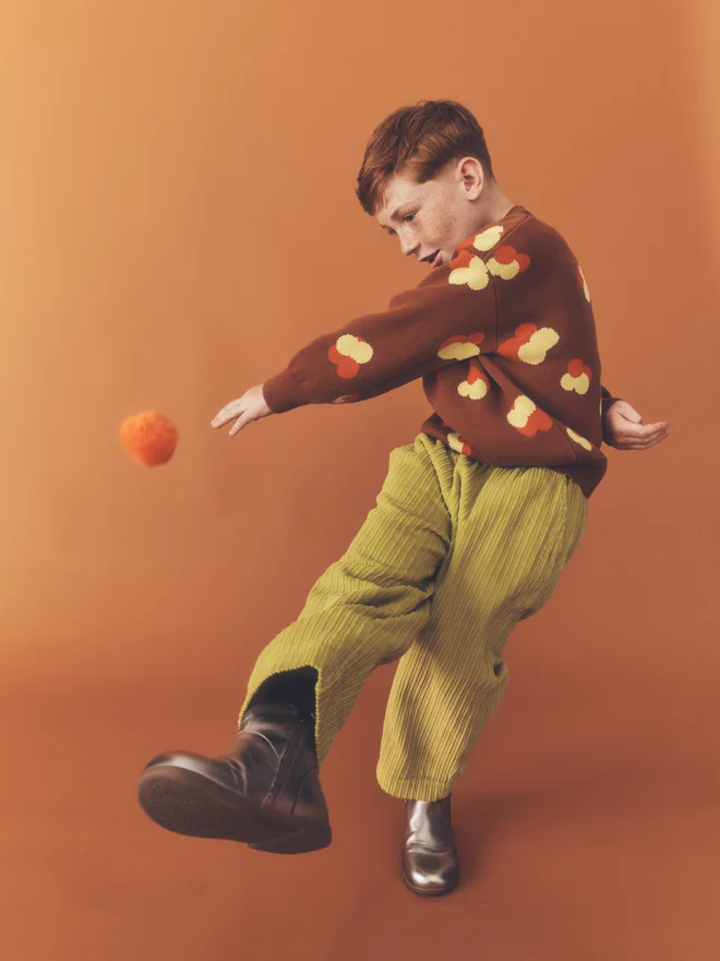 Child kicks an orange pom-pom mid-air against a burnt orange backdrop, wearing a Studio Koter chestnut floral intarsia jumper and chartreuse corduroy trousers.