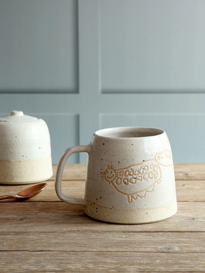 Custom Child's Drawing Stoneware mug with drawing, glazed in our off-white glaze. The mug is sat on a wooden surface with a sugar pot in the background