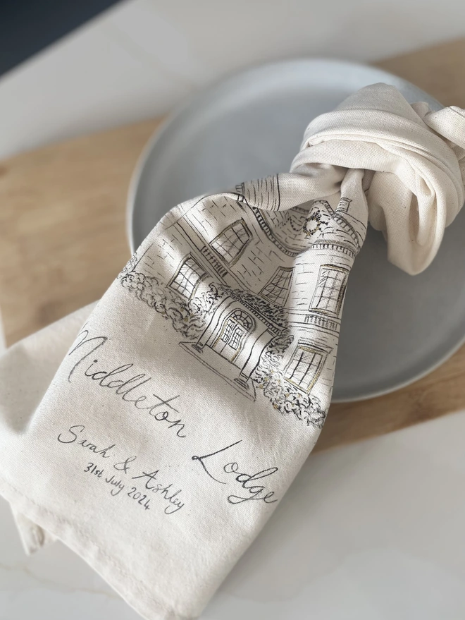 Knotted tea towel with a hand painted venue illustration on a plate with an oak chopping board