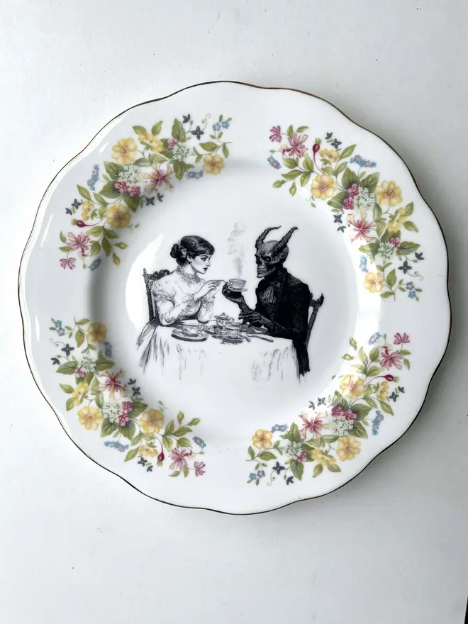 a vintage plate with an ornate edge, ina vintage plate with an ornate edge, in the centre is a vintage illustration of a victorian lady drinking tea with the devil the centre is a vintage illustration of a victorian lady drinking tea with the devil