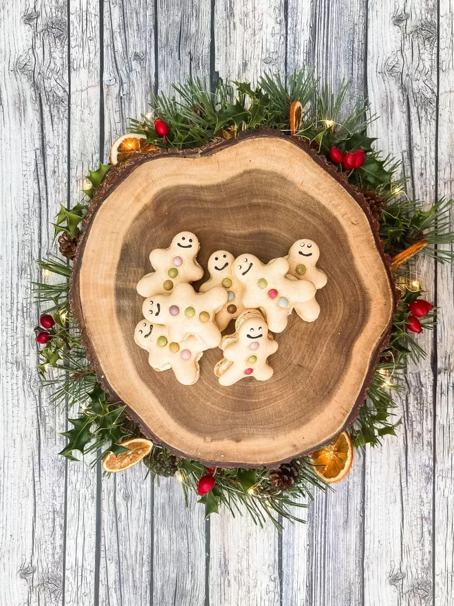 several gingerbread men macarons piled on a wooden plate surrounded by a holly wreath on ash wood background
