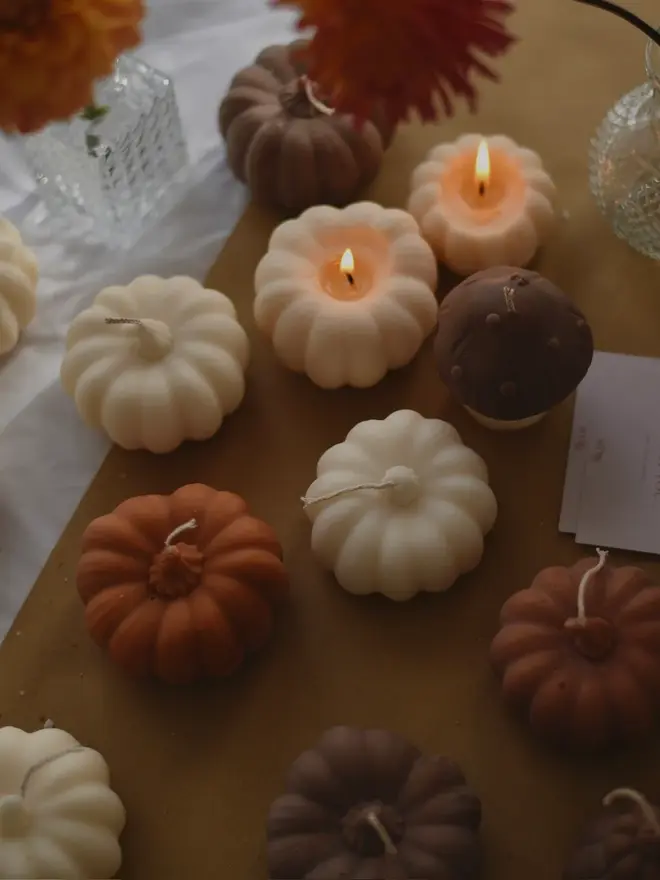 Medium Rust Wax Pumpkin Candle. A collection of pumpkin-shaped candles in cream, terracotta and chocolate brown, with two lit, styled on kraft paper beside orange dahlias, glass vases and thank-you cards. The handmade sculptural candles make a cosy centrepiece for autumn tables, Halloween and Thanksgiving.