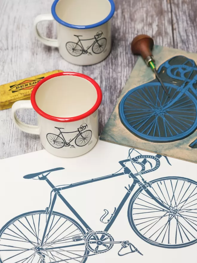 Picture of 2 Cream Enamel Mugs with a bicycle design etched onto it, taken from an original Lino Print. 1 has a blue rim, and the other has a red rim.